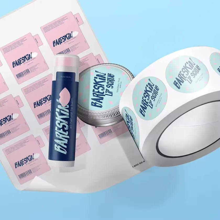 Roll of printed lip balm labels unwinding beside a labelled blue lip balm tube and a lip scrub jar