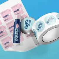 View: Roll of printed lip balm labels unwinding beside a labelled blue tube and a lip scrub jar