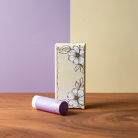 View: Cream lip balm carton with floral line artwork standing beside a lilac lip balm tube on wood