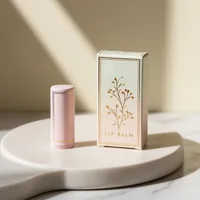 View: Foil-stamped cream lip balm carton standing beside a matching pink lip balm tube on marble