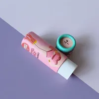 View: Pink illustrated paper lip balm tube lying on its side with its cap off, next to a mint green cap