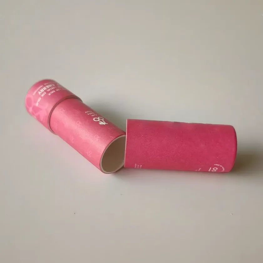 Two magenta printed paper lip balm tubes lying on a grey surface, one with its cap removed